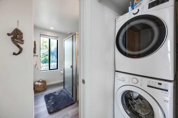 Un cuarto de lavandería con lavadora y secadora apiladas, una puerta de cristal que da a un pasillo, y una cesta tejida en el suelo.