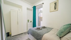 Um quarto com cama, janela com cortinas, guarda-roupa e uma pequena mesa de cabeceira com abajur.