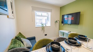 A dining area with a table set for four, a television mounted on the wall, and a window with blinds.