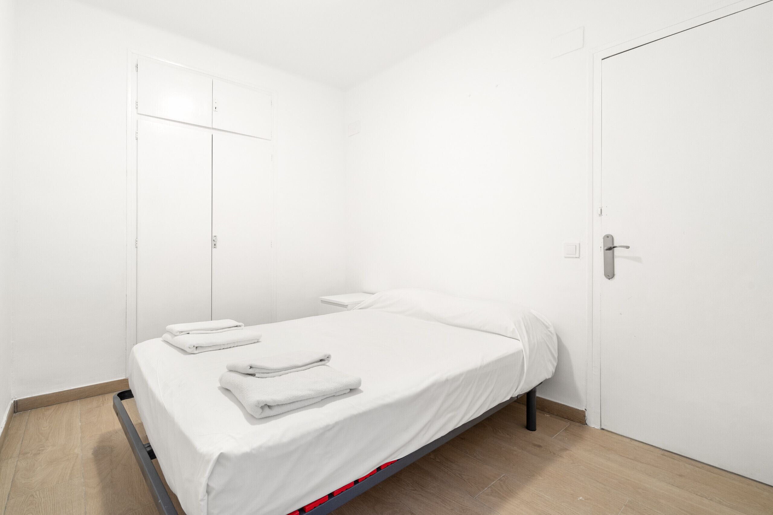Una cama individual con ropa blanca de cama y una mesita de noche pequeña, una puerta blanca y un suelo de madera.