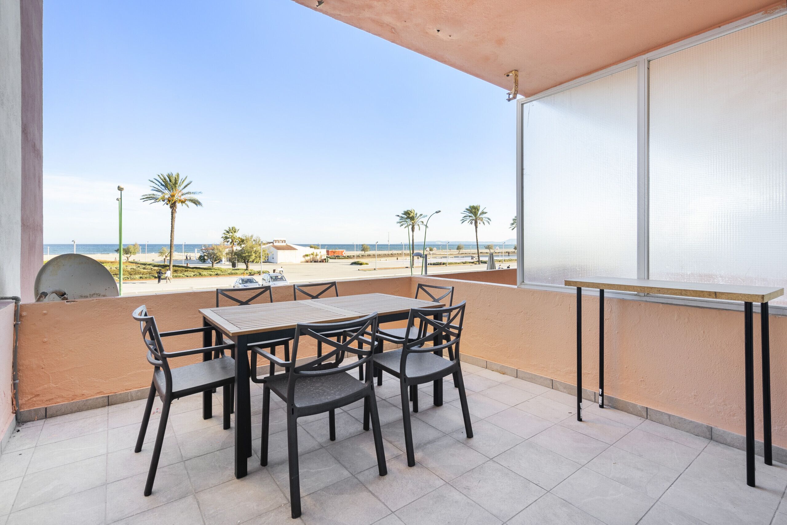 A balcony with a table and chairs, overlooking a beach with palm trees and a clear sky.