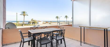 A balcony with a table and chairs, overlooking a beach with palm trees and a clear sky.