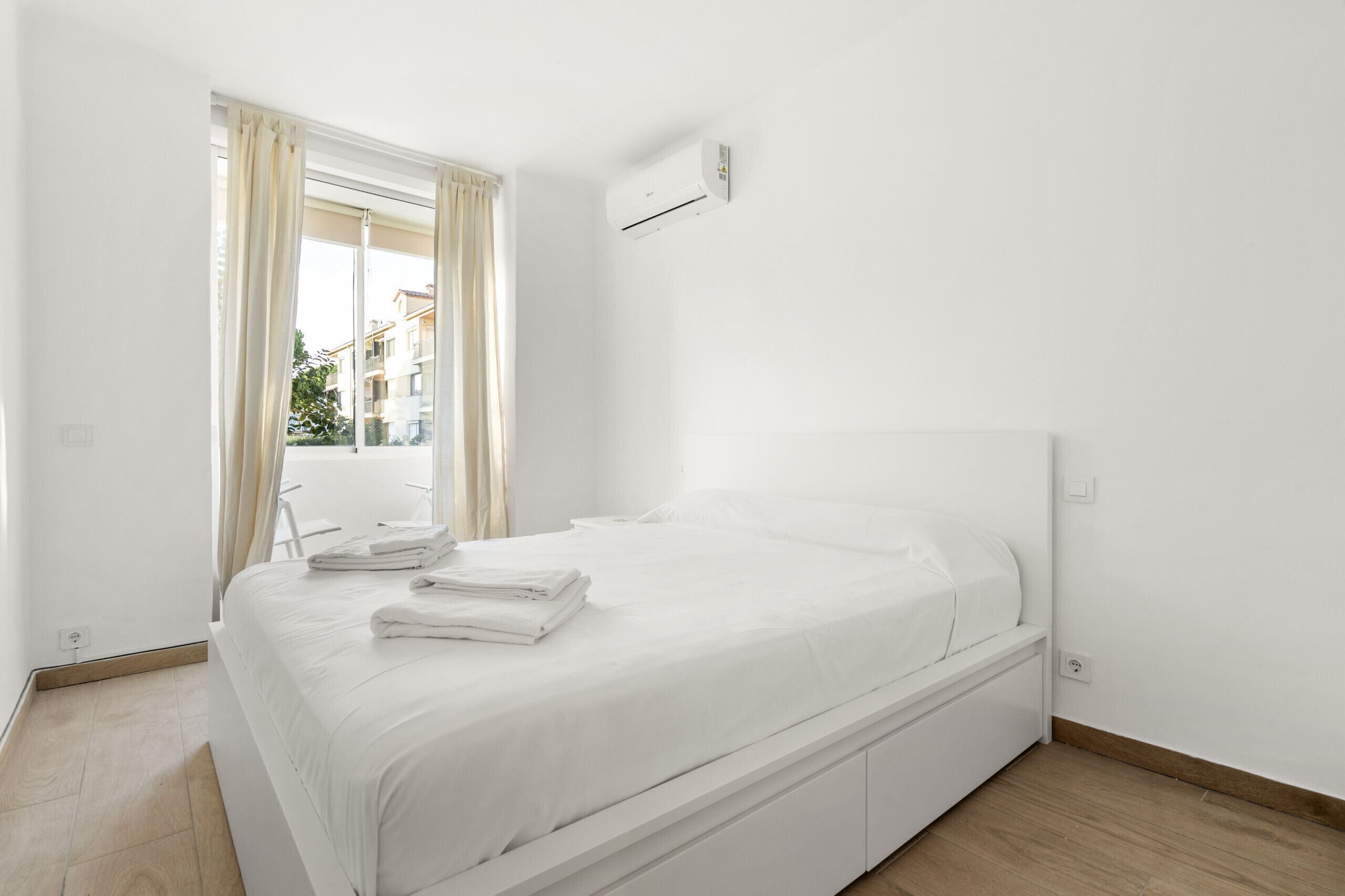A clean, white hotel room with a bed, a window with curtains, and a wall-mounted air conditioning unit.