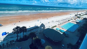 A beach view with a pool area, palm trees, and beach chairs.