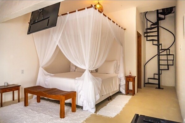 Een slaapkamer met een hemelbed, een houten bankje, een klein nachtkastje, een witte vloerkleed en een spiltrap op de achtergrond.
