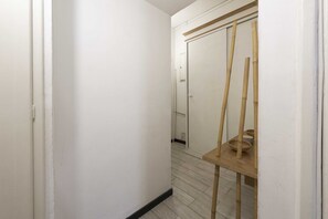Um corredor estreito com paredes brancas e chão de madeira, que dá para um quarto com porta e uma pequena mesa com cestos.