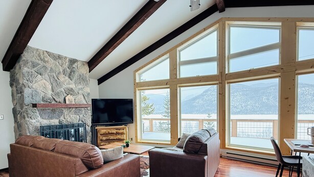 Amplia sala de estar con chimenea de piedra, vigas de madera y ventanas amplias que ofrecen vistas a montañas nevadas.