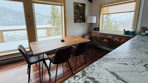 Un comedor con mesa y sillas de madera, ventanas amplias con vistas a montañas nevadas y una cocina moderna con encimera de mármol.