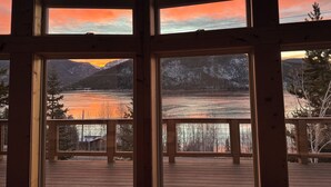 Una cabaña con amplias ventanas que ofrecen vistas a un lago y montañas durante el atardecer.