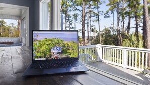 Ein Laptop auf einem Holztisch mit Blick auf ein Haus, das von Bäumen und einem Gewässer umgeben ist.