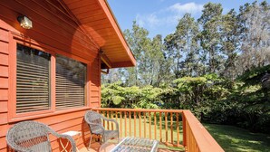 Un patio in legno con mobili in vimini e un tavolo in vetro, circondato da vegetazione rigogliosa e alberi.