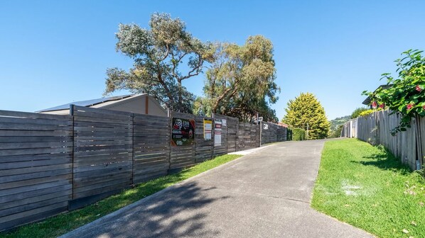 A gated driveway leading to a building with a sloped roof, surrounded by a wooden fence and greenery.