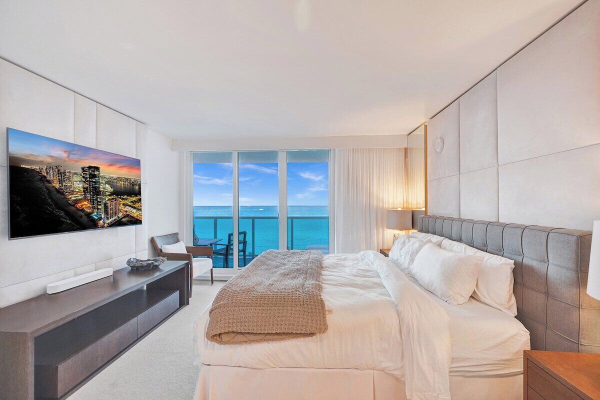Une chambre d’hôtel moderne dotée d’un grand lit, d’un téléviseur à écran plat fixé au mur, d’un balcon avec vue sur la mer et d’un coin salon avec une table et des chaises.