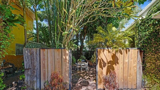A pathway leading to a wooden gate, surrounded by lush greenery and a yellow building. - Luxury Morningside Bungalow Steps to the Park (Miami)