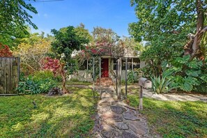 Un sendero en un jardín que lleva a una casa con puerta roja, rodeada de plantas y árboles.