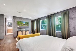 A modern hotel room with a large bed, a television mounted on the wall, and a view of the outdoors through the windows.