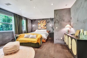 A modern bedroom with a bed, bedside table, lamp, and a view of greenery outside.