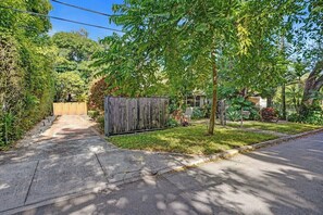 Una entrada de casa con cerramiento de madera, un camino pavimentado y un área verde con árboles.
