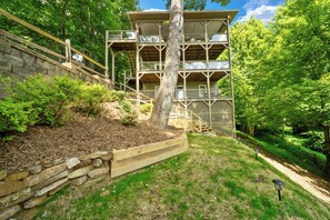 A multi-level wooden cabin with a stone retaining wall, surrounded by a wooded area.