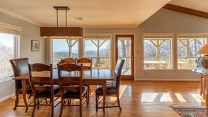 Un espace repas avec une table et des chaises en bois, de grandes fenêtres offrant une vue dégagée et un luminaire suspendu.