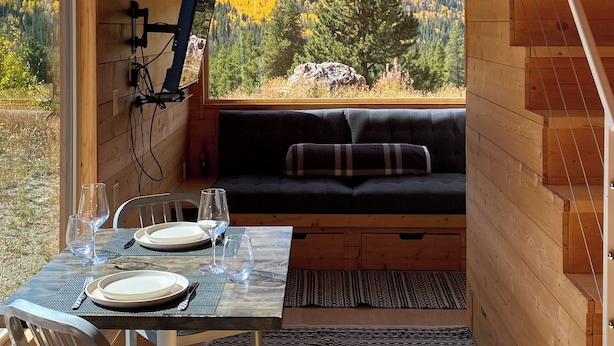 Interior acogedor de una cabaña con comedor, sofá y ventana con vistas a un bosque.