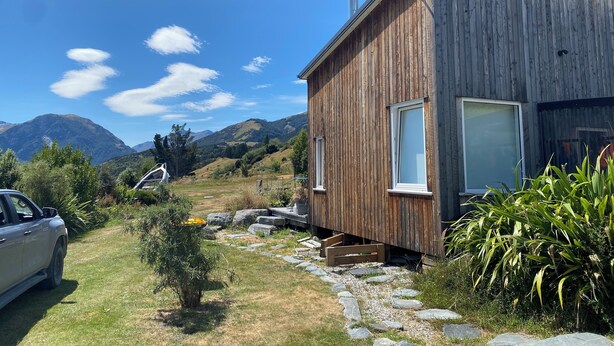 Eine Holzhütte mit einer Satellitenschüssel, ein geparktes Auto und eine gebirgige Landschaft im Hintergrund.