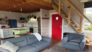 Ein modernes Wohnzimmer mit einem grauen Sofa, einem blauen Sessel und einem Holztisch. Im Hintergrund sind eine rote Tür und eine Holztreppe zu sehen.