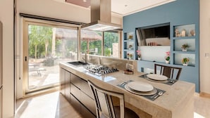 A modern kitchen with a large island, a gas stove, and a sink. There are two bar stools and a dining table set for two. The kitchen has a blue accent wall and a large window overlooking a garden. - Tulum Country Home (Akumal)