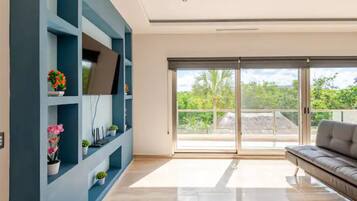 A modern living room with a sofa, a TV on a blue wall, and a balcony with a view of greenery.