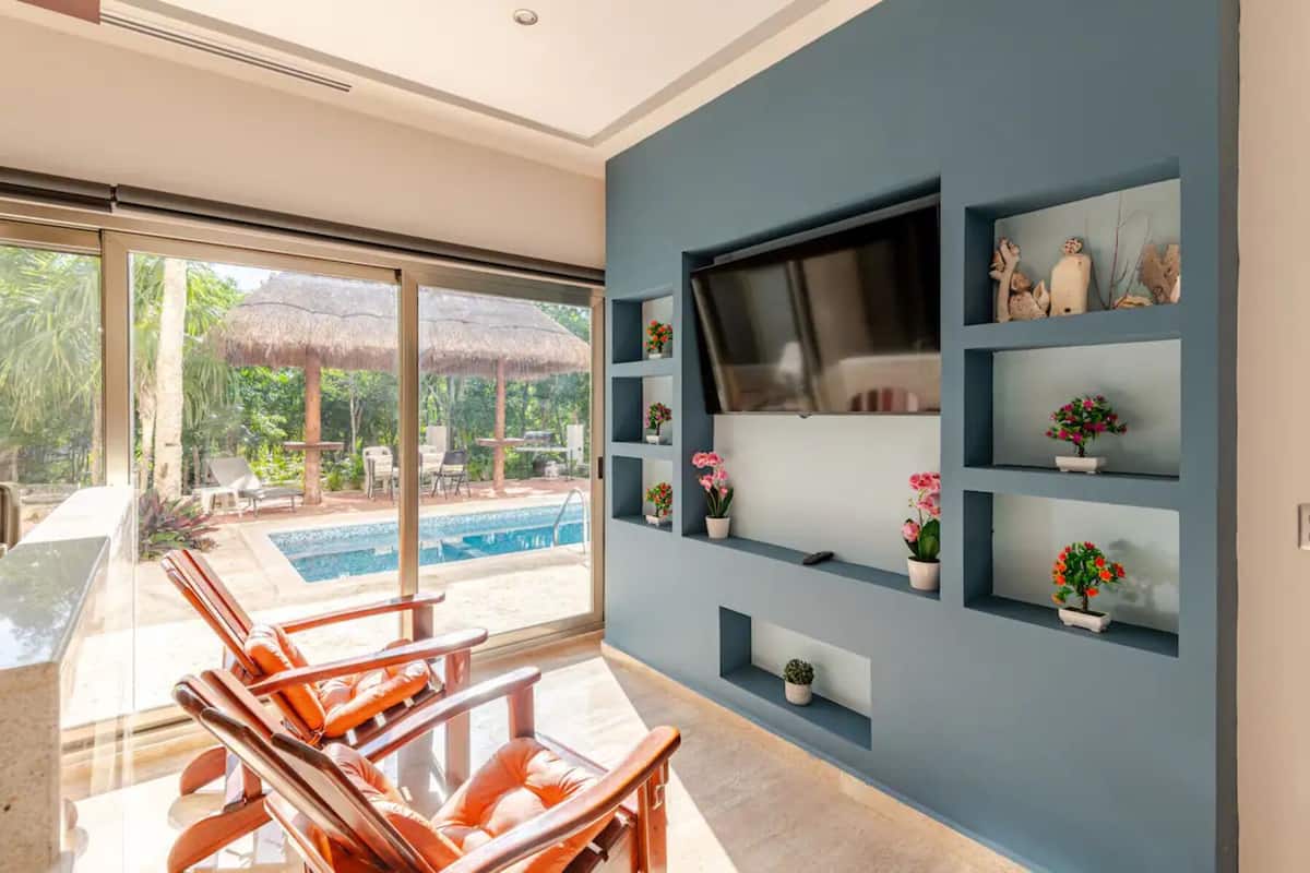 A modern living room with a large TV, built-in shelves with potted plants, and a view of an outdoor pool area.