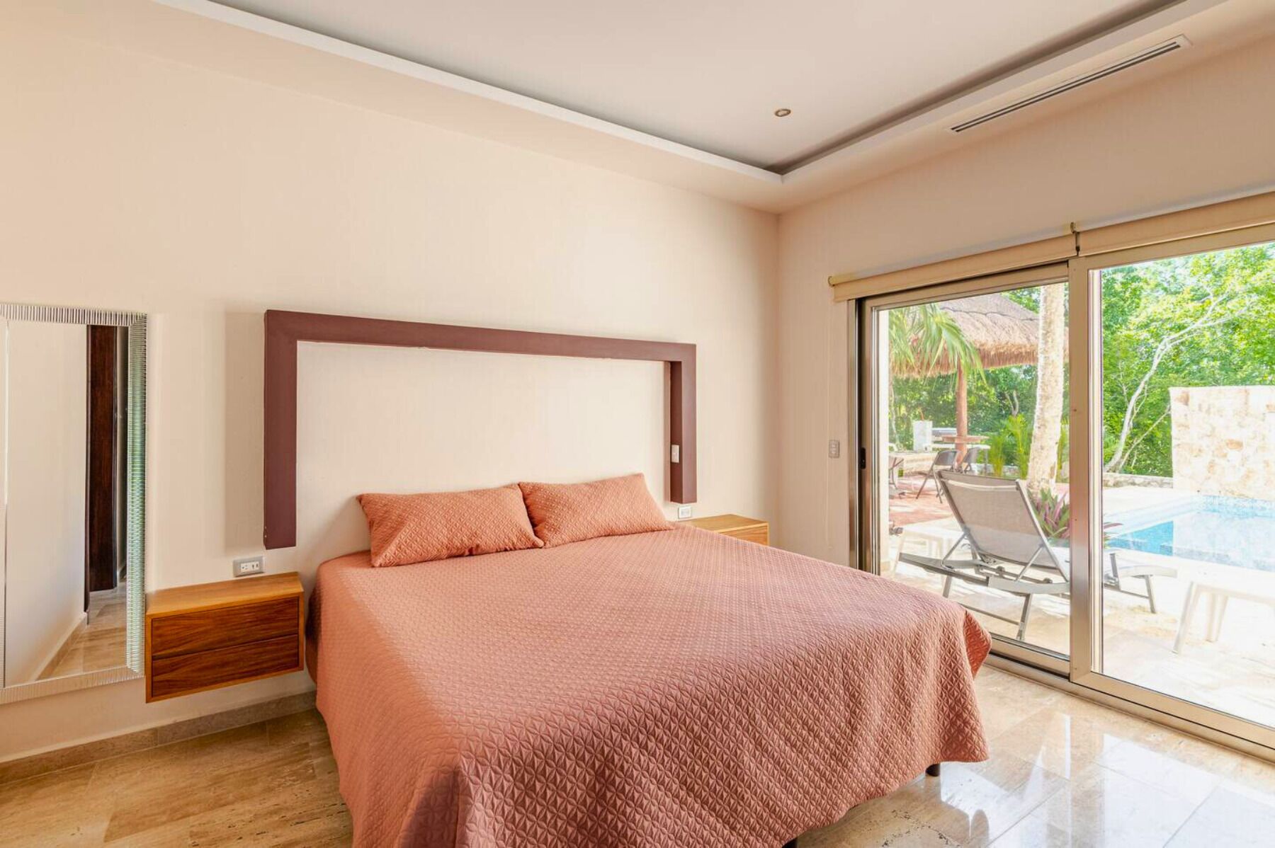 Ein Schlafzimmer mit einem Bett, einem Nachttisch, einer Holzkistenreih und einer großen Schiebetür zu einem Außenbereich mit Pool und Liegestühlen.