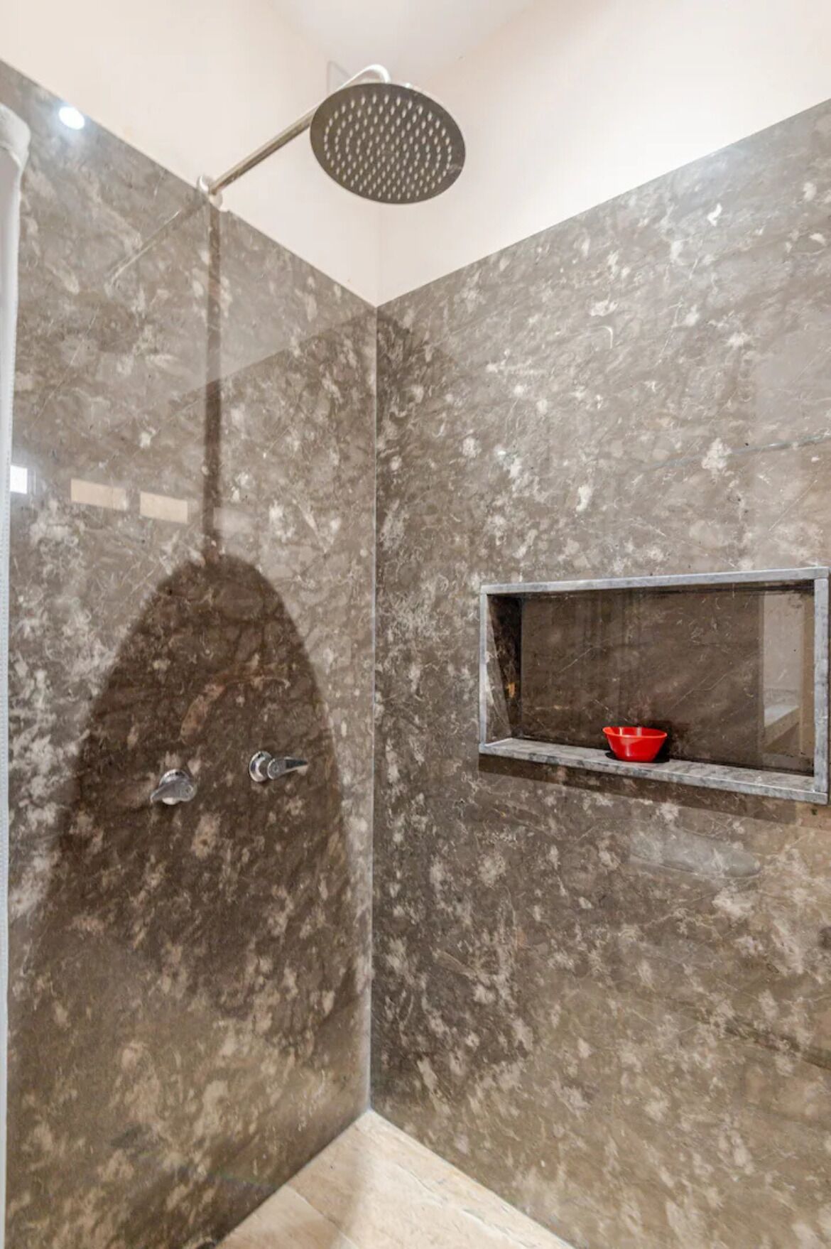 Área de duche com chuveiro de teto, prateleira embutida com uma tigela vermelha e paredes de mármore.