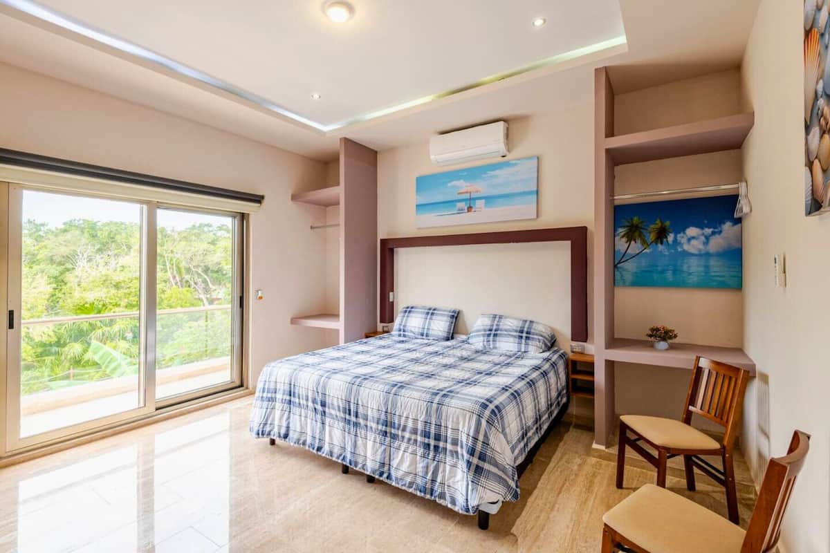 A bedroom with a large bed, a chair, a painting, and a view of greenery through a sliding door.