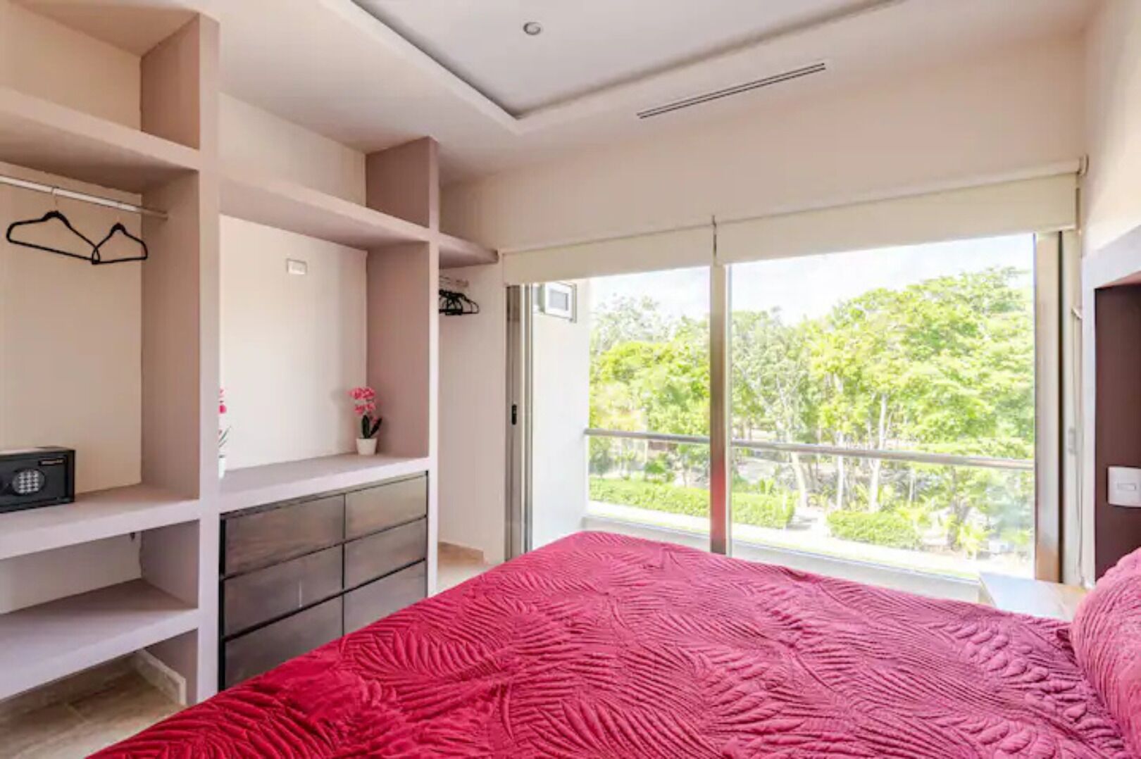 Un dormitorio con un ventanal grande, una cama con colcha roja y estanterías empotradas.
