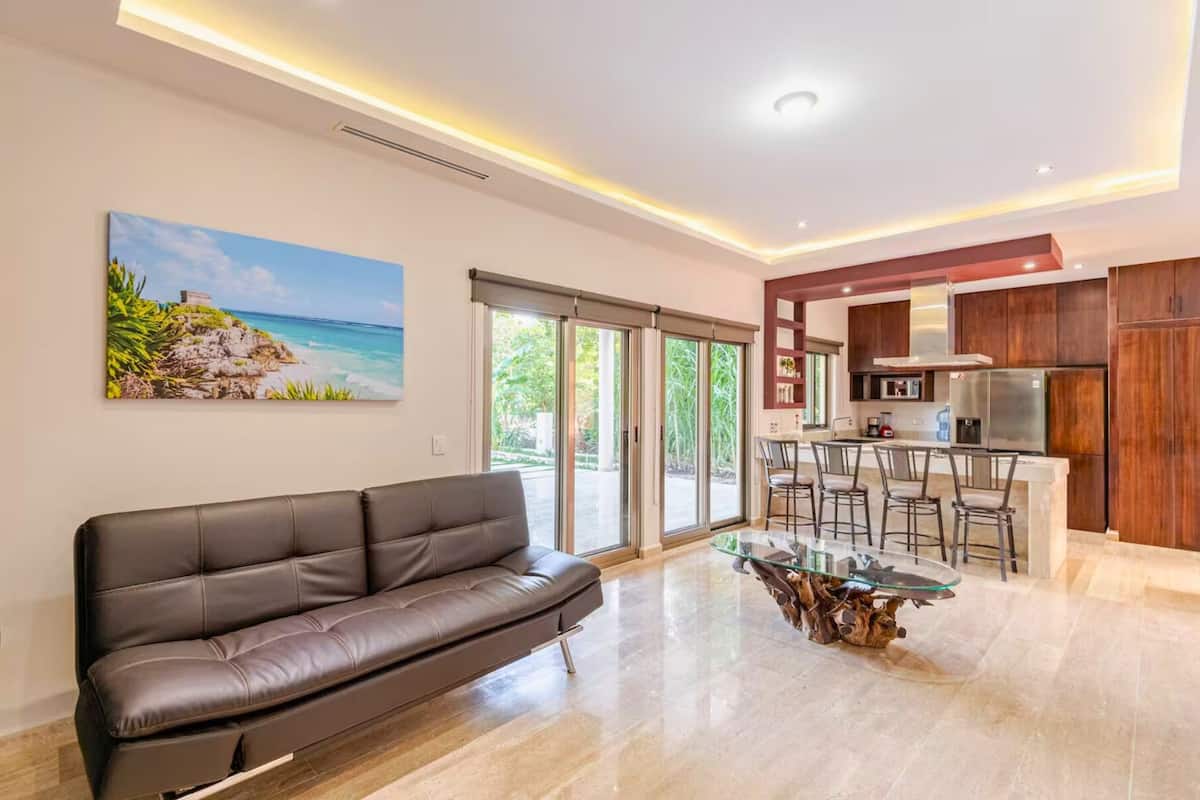 A modern living room with a leather sofa, a glass coffee table, and a kitchen area with wooden cabinets and a stainless steel refrigerator.