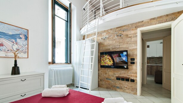 A modern hotel room with a large bed, a TV mounted on a brick wall, a window with blinds, and a view of the kitchen. - San Giusto 21 Old Town - Lecce Selection (Lecce)