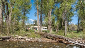 Une rivière aux eaux claires, bordée de grands arbres, avec une maison qui se profile à l’arrière-plan.