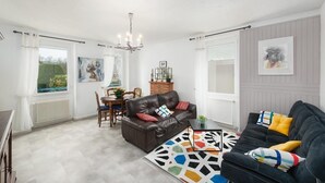 A modern living room with a brown leather sofa, a black sofa, and a colorful geometric rug. There is a dining table with chairs, a chandelier, and artwork on the walls.