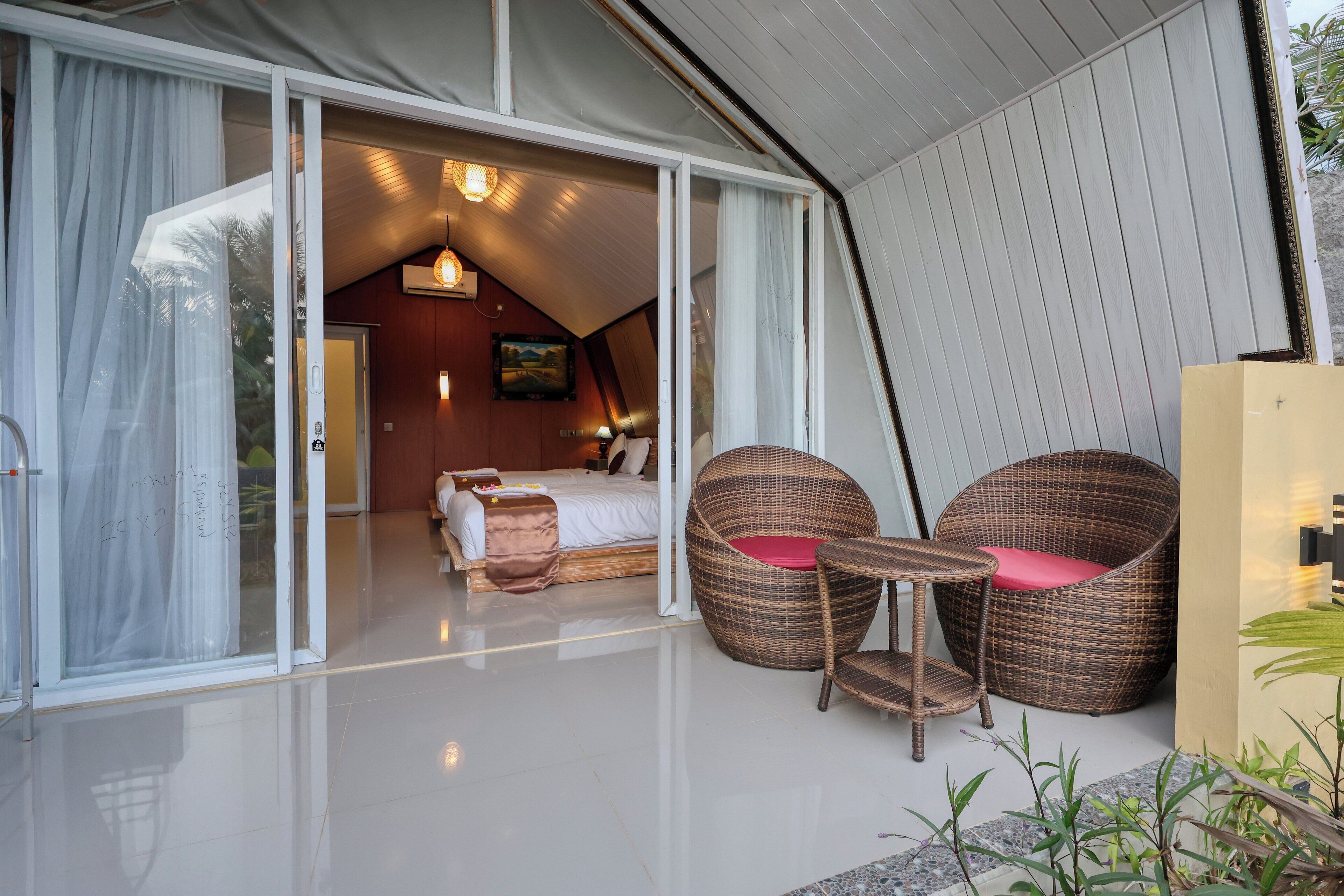 A modern hotel room with a large sliding glass door leading to a balcony, a bed with a white duvet, and two wicker chairs with red cushions.