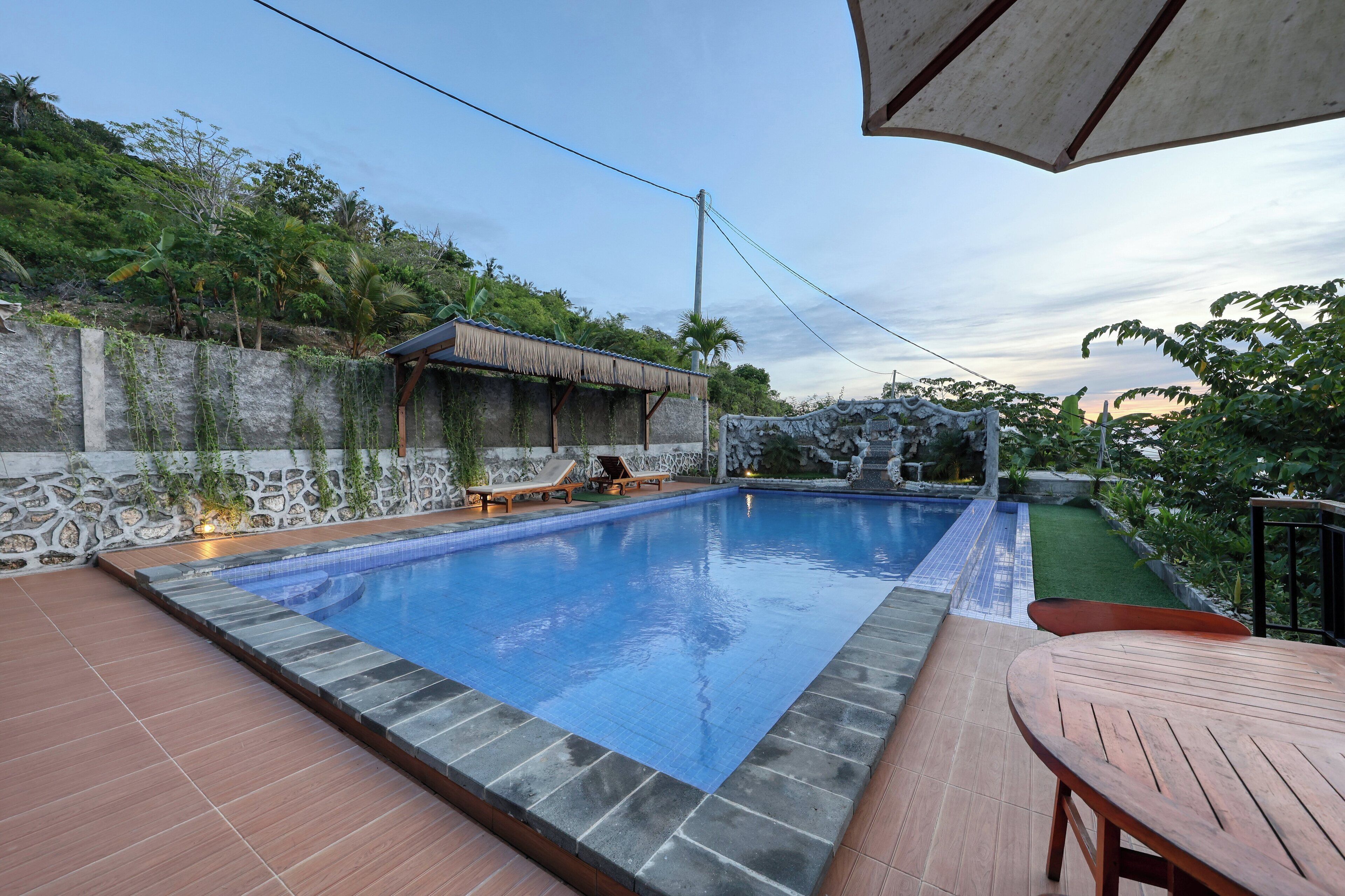Une piscine bordée de pierres, une terrasse en bois et une structure surmontée d’un toit de chaume.