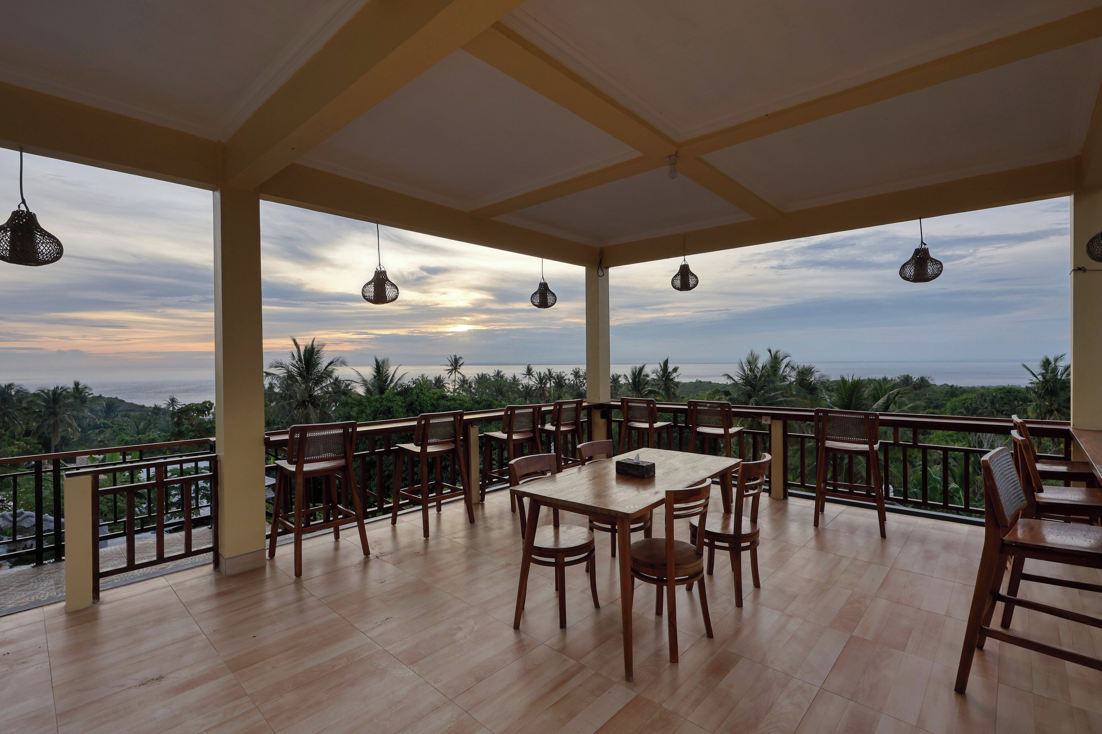 A covered outdoor seating area with wooden tables and chairs, overlooking a scenic view of the ocean and palm trees.