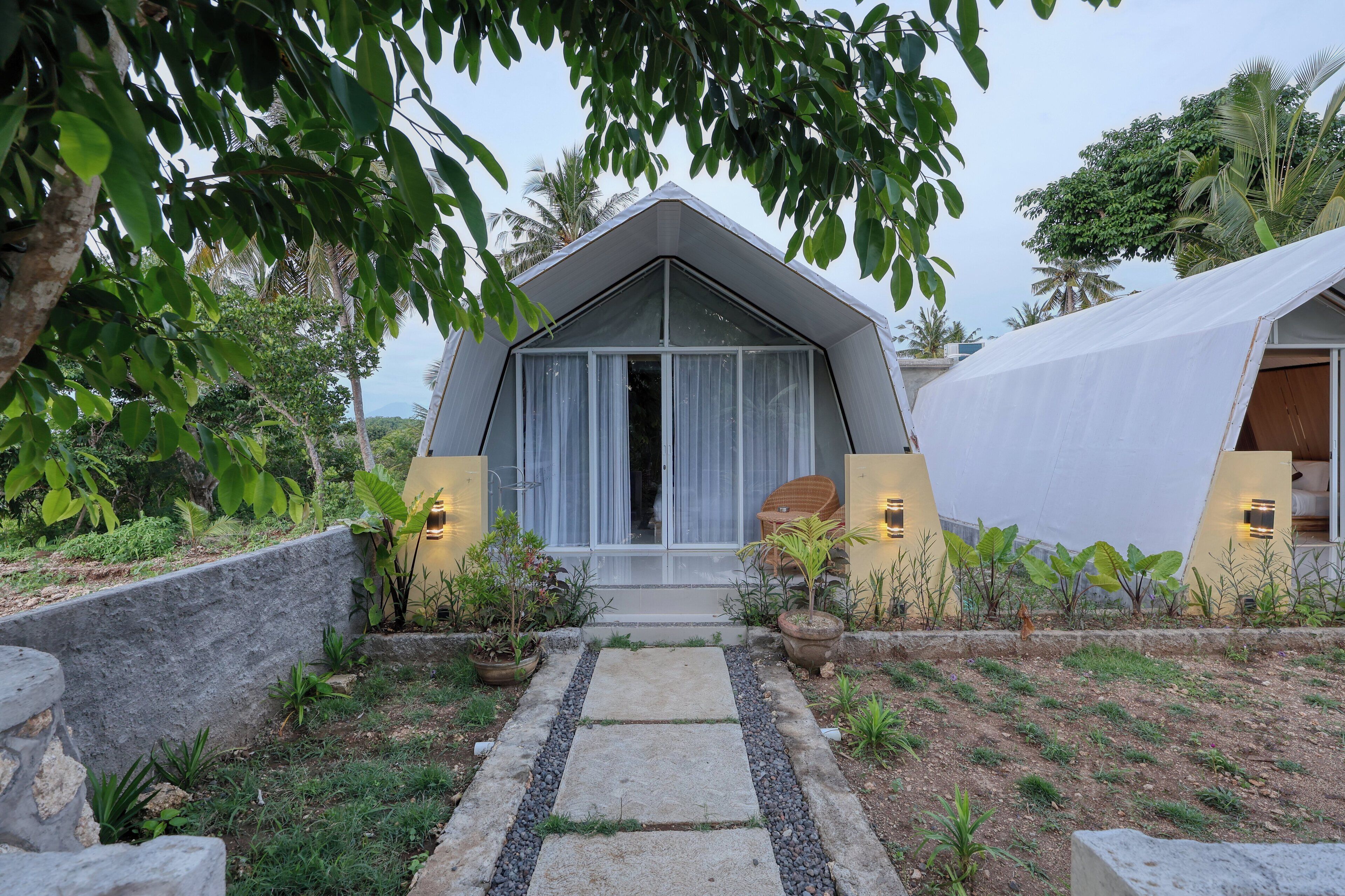 Un edificio moderno de forma triangular con puerta y ventanas de vidrio, rodeado por un jardín con plantas en macetas y un sendero de piedra.