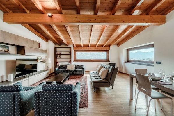 A modern living room with a TV, sofas, a dining table, and a large window with a mountain view.
