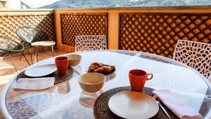 Una mesa redonda de vidrio con platos, tazones y tazas, rodeada de sillas de mimbre, ubicada en una terraza con vistas a las montañas.