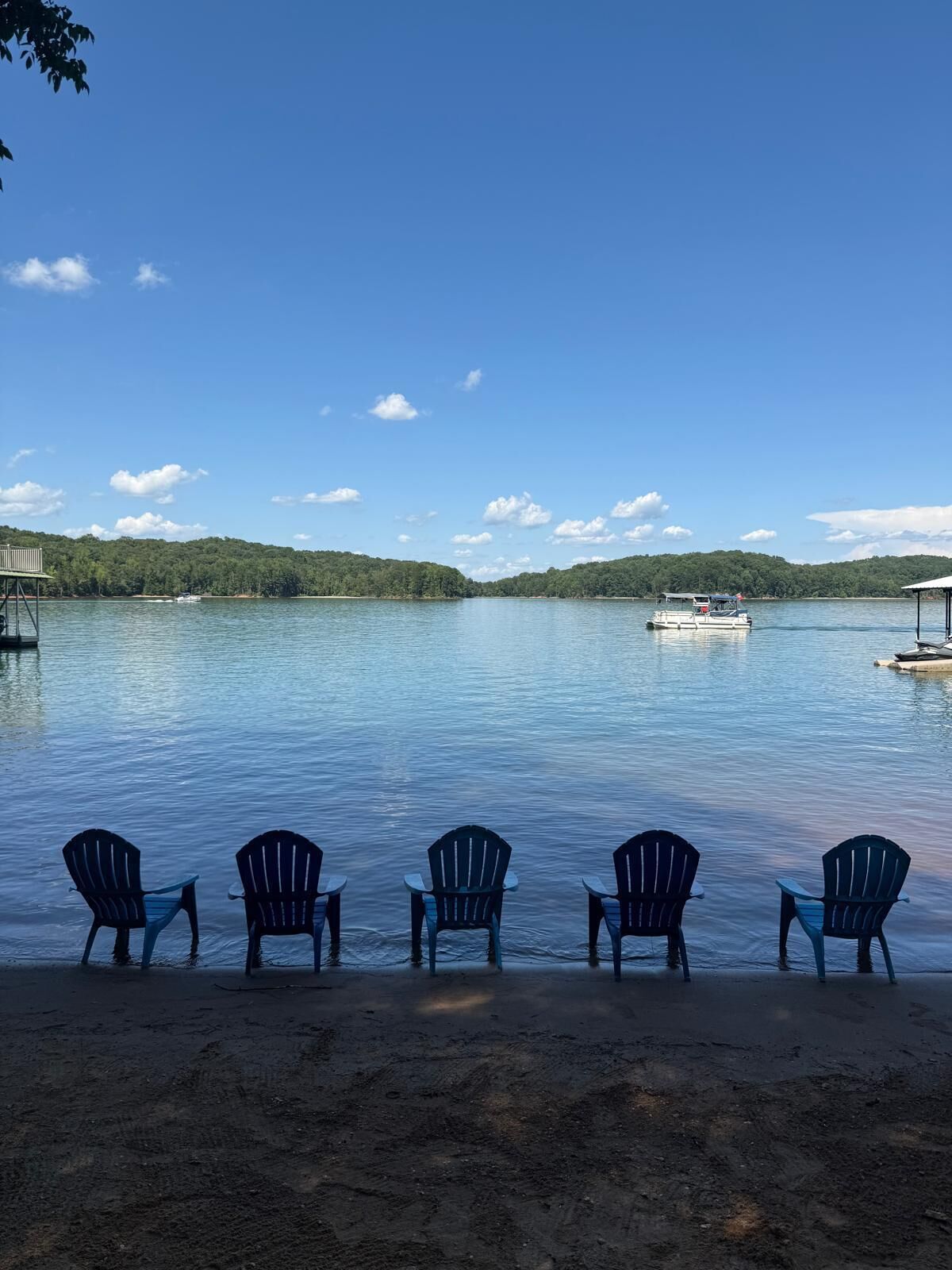 A lakeside view with five Adirondack chairs facing the water, a dock with a boat, and a forested hill in the background.