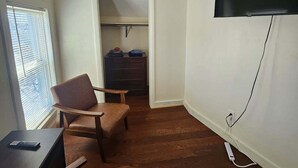 A room with a wooden floor, a TV mounted on the wall, a chair, a desk, and a window with blinds. - Nice apartment near Whole Foods, Brown University, Miriam Hospital, Downtown (Providence)