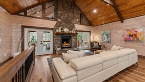 A log cabin interior with a stone fireplace, wooden ceiling beams, and a large white sofa. - Cardinal Lodge - Elevated Luxury, Minutes From Fun (Mineral Bluff)