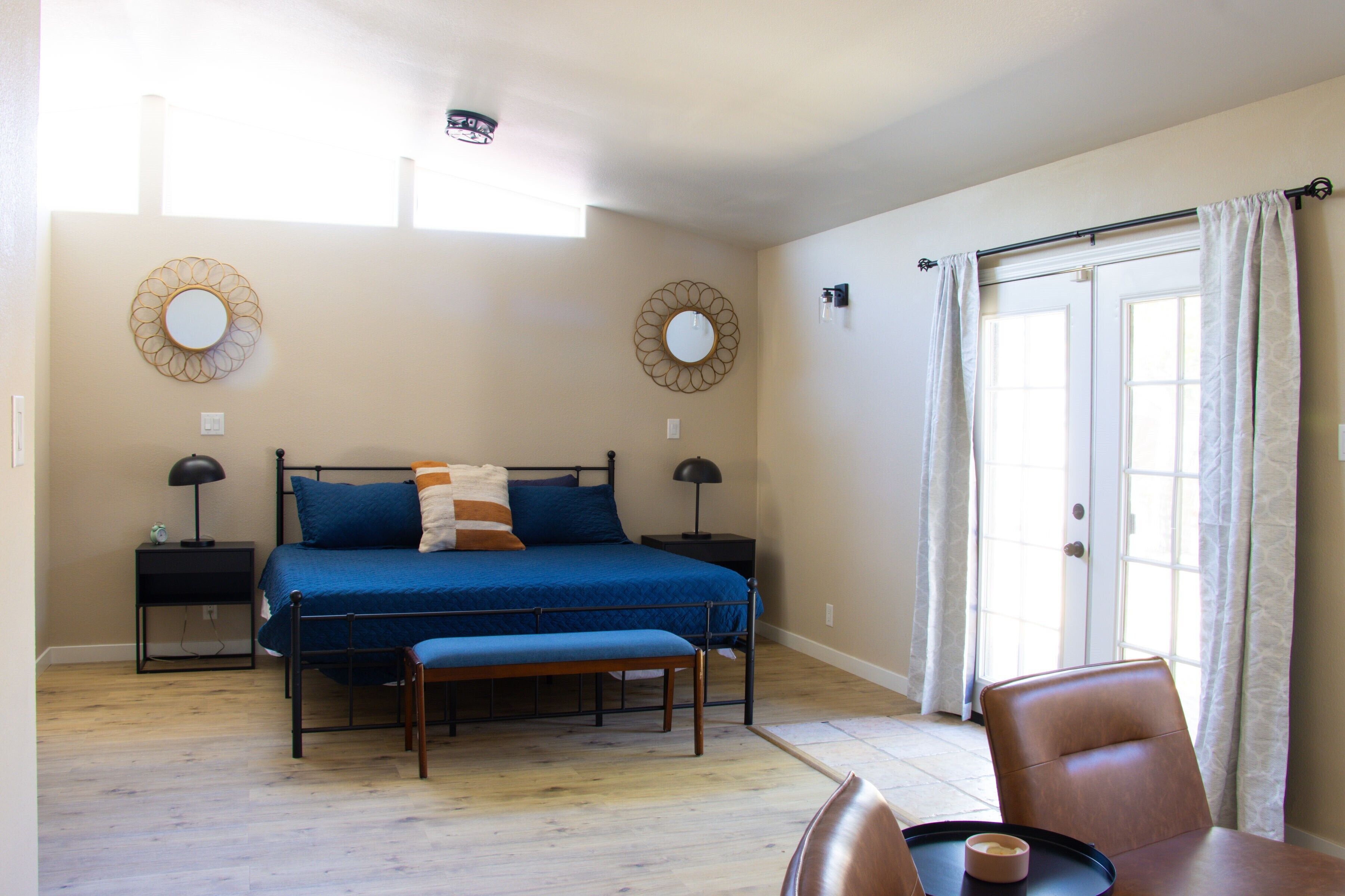 A bedroom with a bed, bedside tables, a bench, a dining table, and a view of the outdoors through a door.