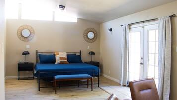 A bedroom with a bed, bedside tables, a bench, a dining table, and a view of the outdoors through a door.
