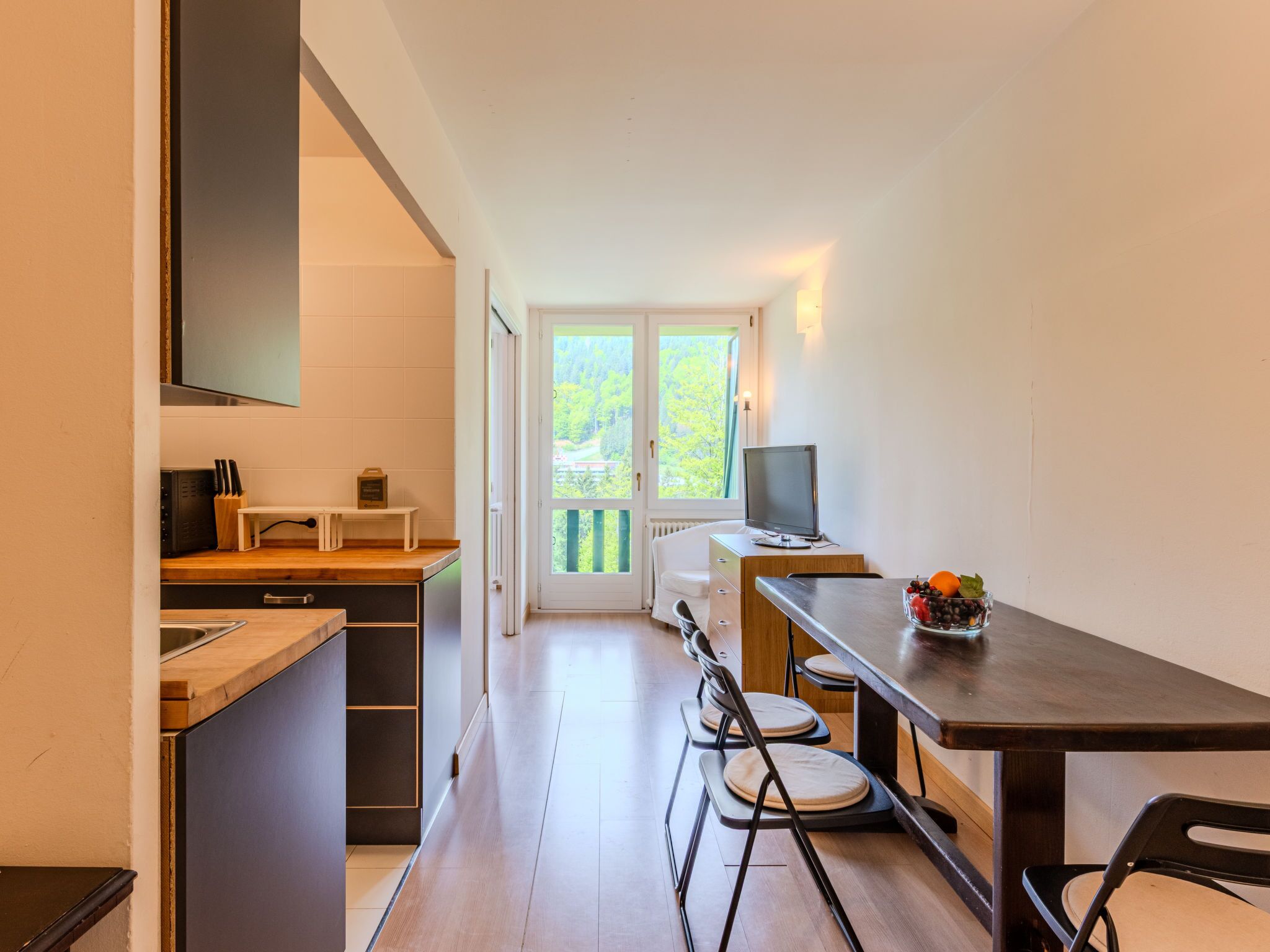 Una cocina compacta con microondas, tostadora y una pequeña mesa con un tazón de fruta. Se ve una ventana con vista a árboles y una puerta que da a un balcón.